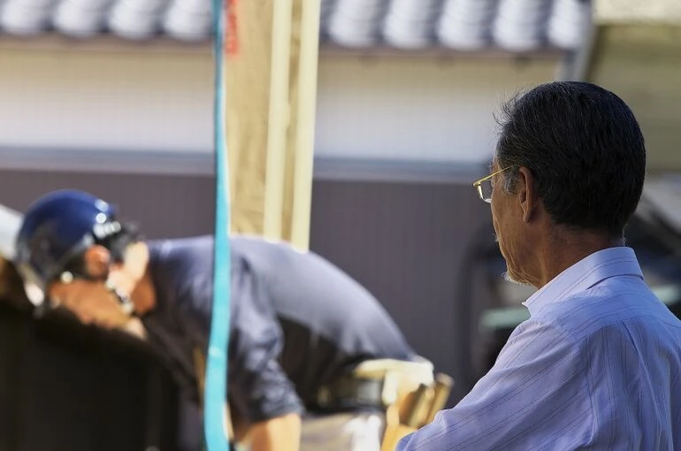 現場で若い大工の仕事を見守る職人経験者の姿。技術継承の重要性と大工不足の現実を象徴する