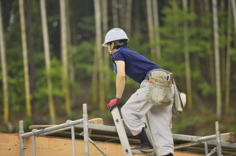竹林を背景に、木材を担いで足場の上で作業する大工職人の後ろ姿。自然の中で木と向き合いながら家づくりを進める様子