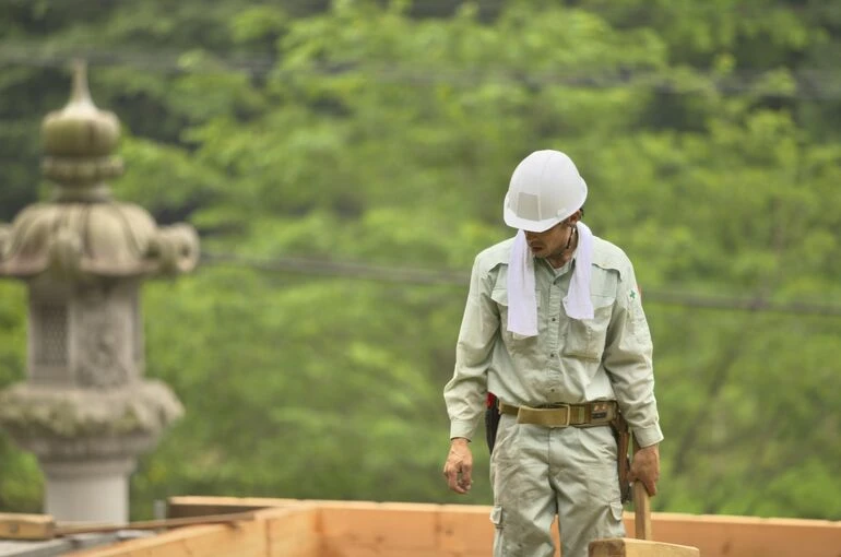 木の家の土台の上で道具を手に、作業に集中する大工職人の姿。背景の緑と寺院の灯籠が、伝統的な家づくりの雰囲気を引き立てている。