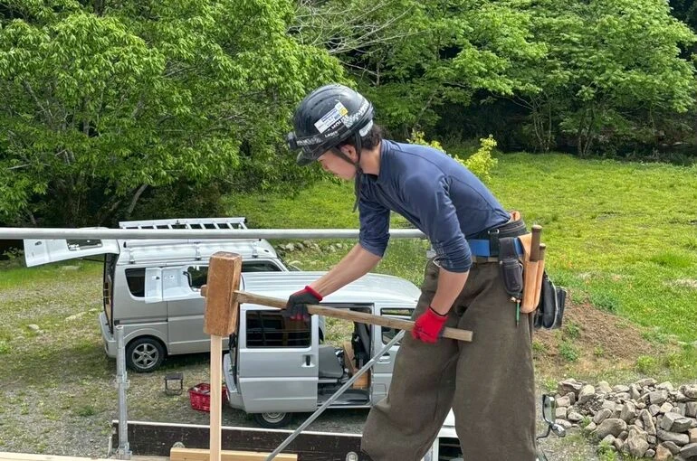 建て方作業中、梁の上で木槌を使い仕口を締め込む若い大工職人。山間の現場で伝統的な木組み施工を行う様子が伝わる
